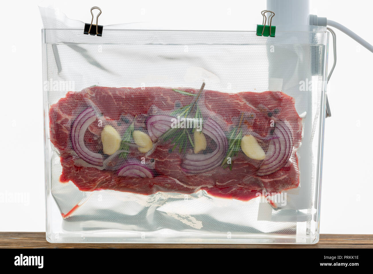 Portion of flat iron beef steak sous-vide cooking suspended in a clear plastic bag in hot water with a spirg of fresh rosemary and onions to flavor Stock Photo