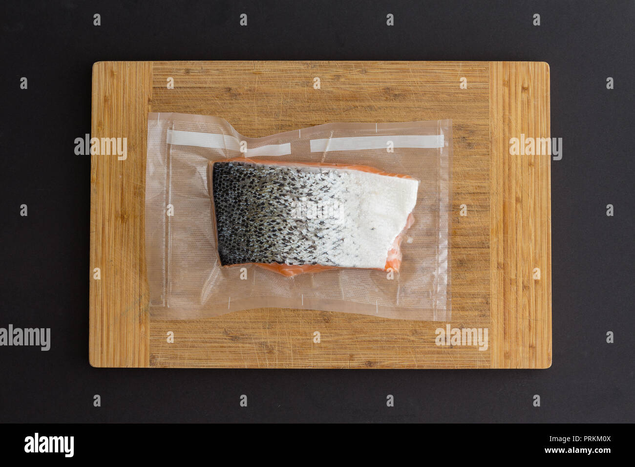 Fillet of fresh Atlantic salmon vacuum packed for freezing in clear plastic lying on a bamboo board over black in a top down view Stock Photo