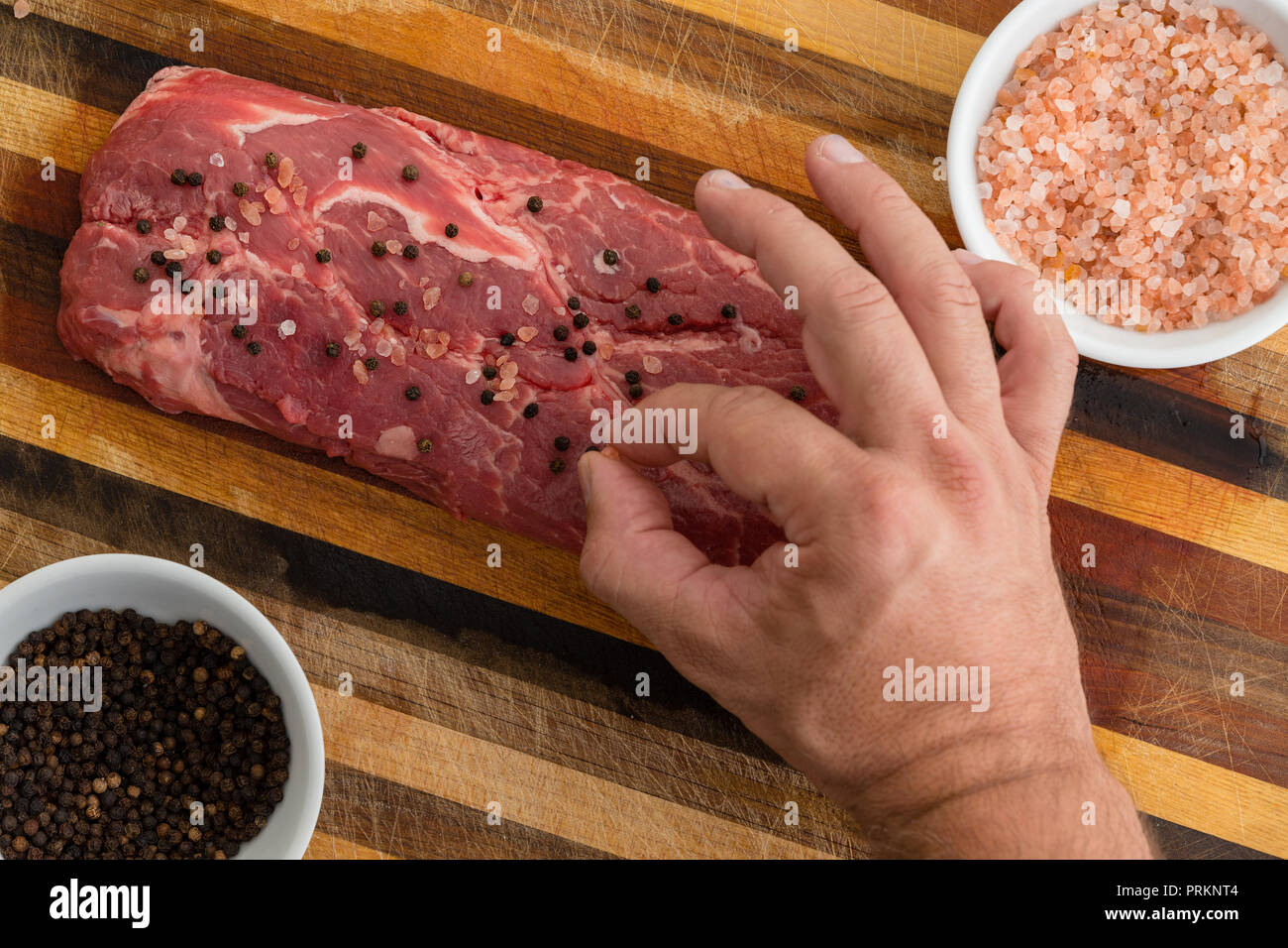 Man dropping seasoning on slab of red meat sitting on striped wooden table next to bowls of pepper and salt Stock Photo