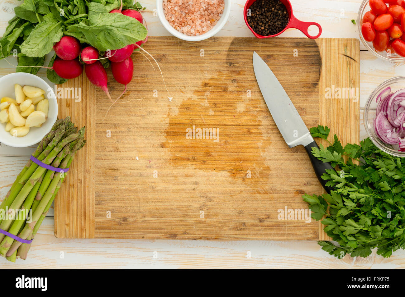 Overhead view of wooden cutting board surrounded by assorted vegetables including onion and asparagus with knife Stock Photo