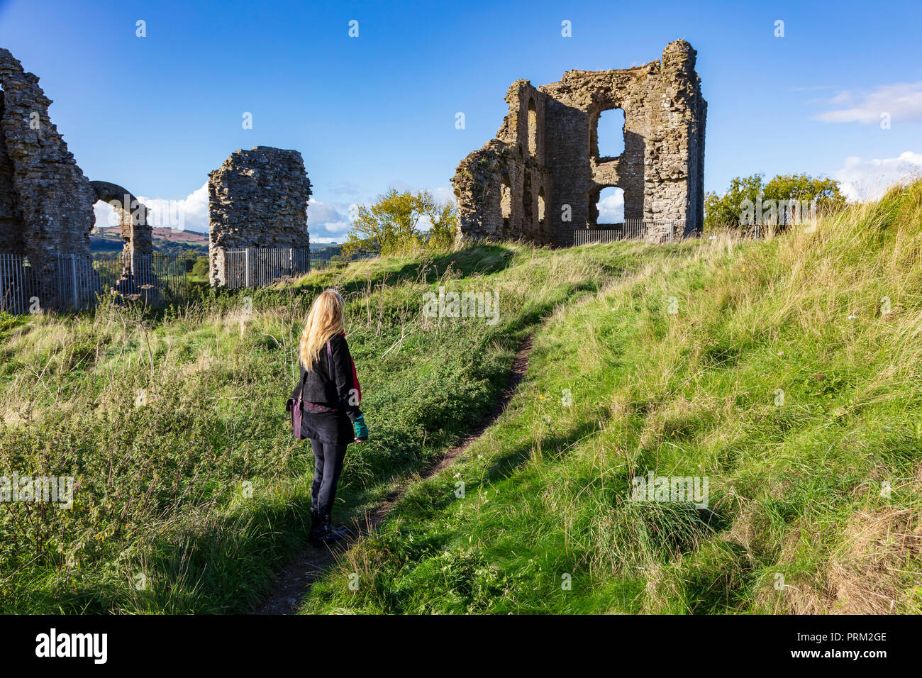 A blonde woman stands on a narrow path and looks at the remains of Clun Castle  on top of a small hill, the area is quiet at the start of Autumn on a sunny day, Shropshire, UK Stock Photo