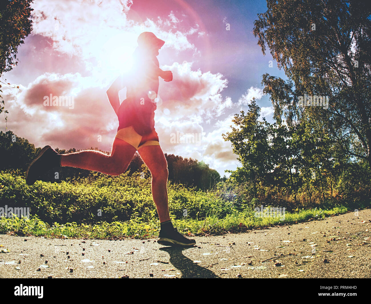 Jogging tall sports man in trees shadows with sun light behind him while wearing black yellow shorts and blue jogging attire Stock Photo