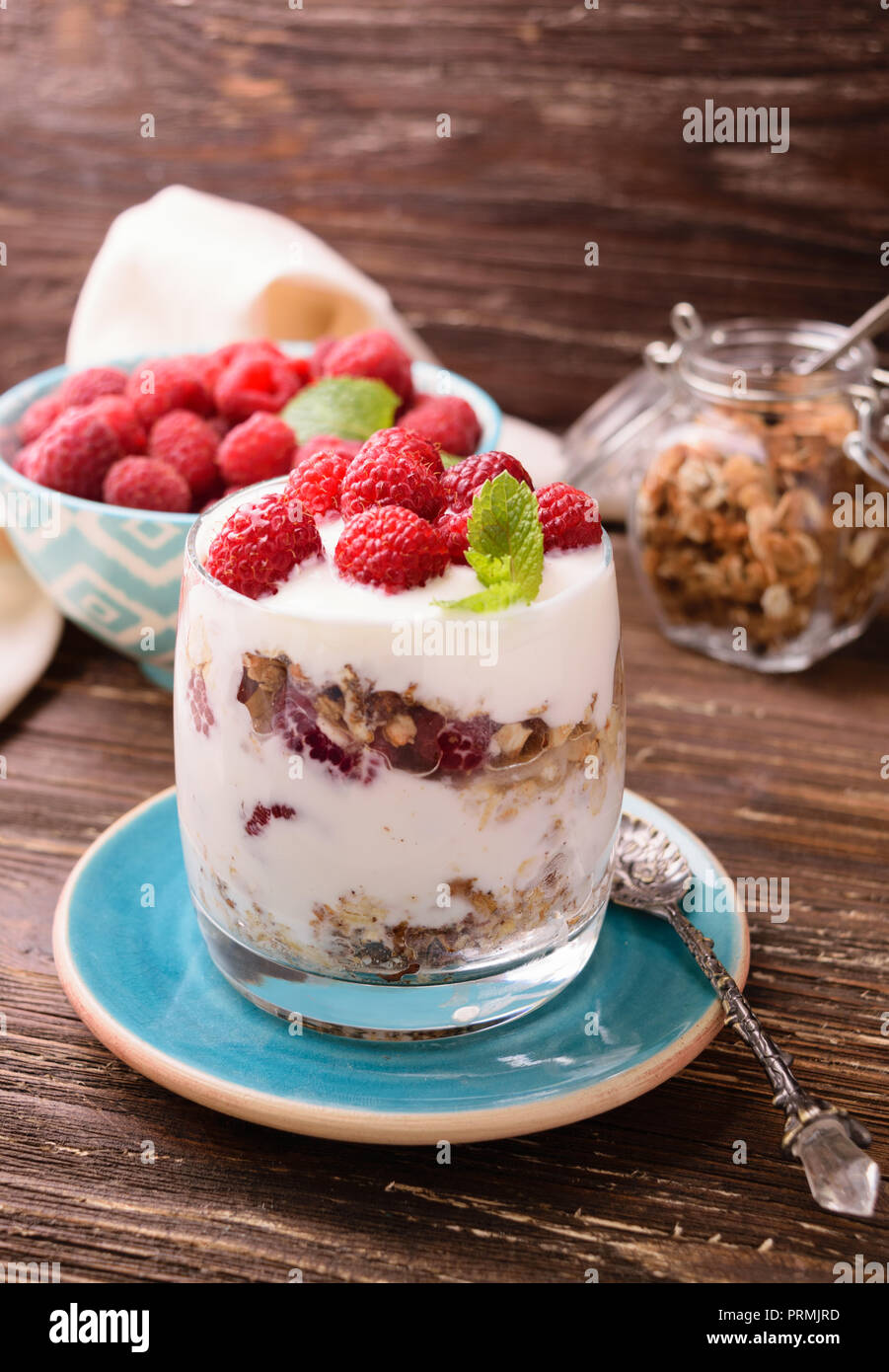 Layered yogurt with granola and raspberries. In a glass, decorated with mint leaves. On dark rustic wooden table background. Healthy food concept. Stock Photo