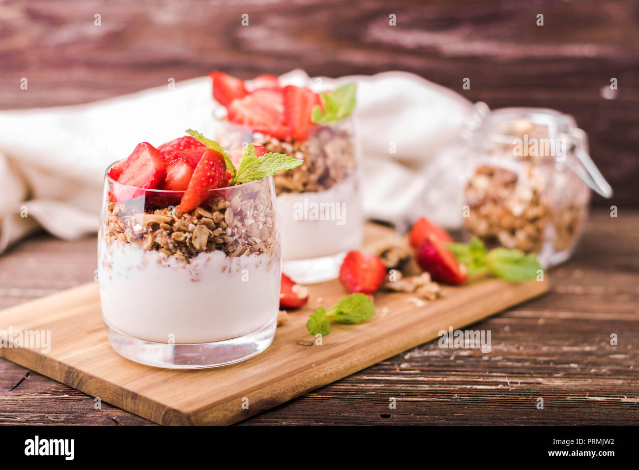 Yogurt parfait in glasses with granola, strawberries and mint leaves. On wooden board. Healthy breakfast concept. Rustic wooden table background. Stock Photo