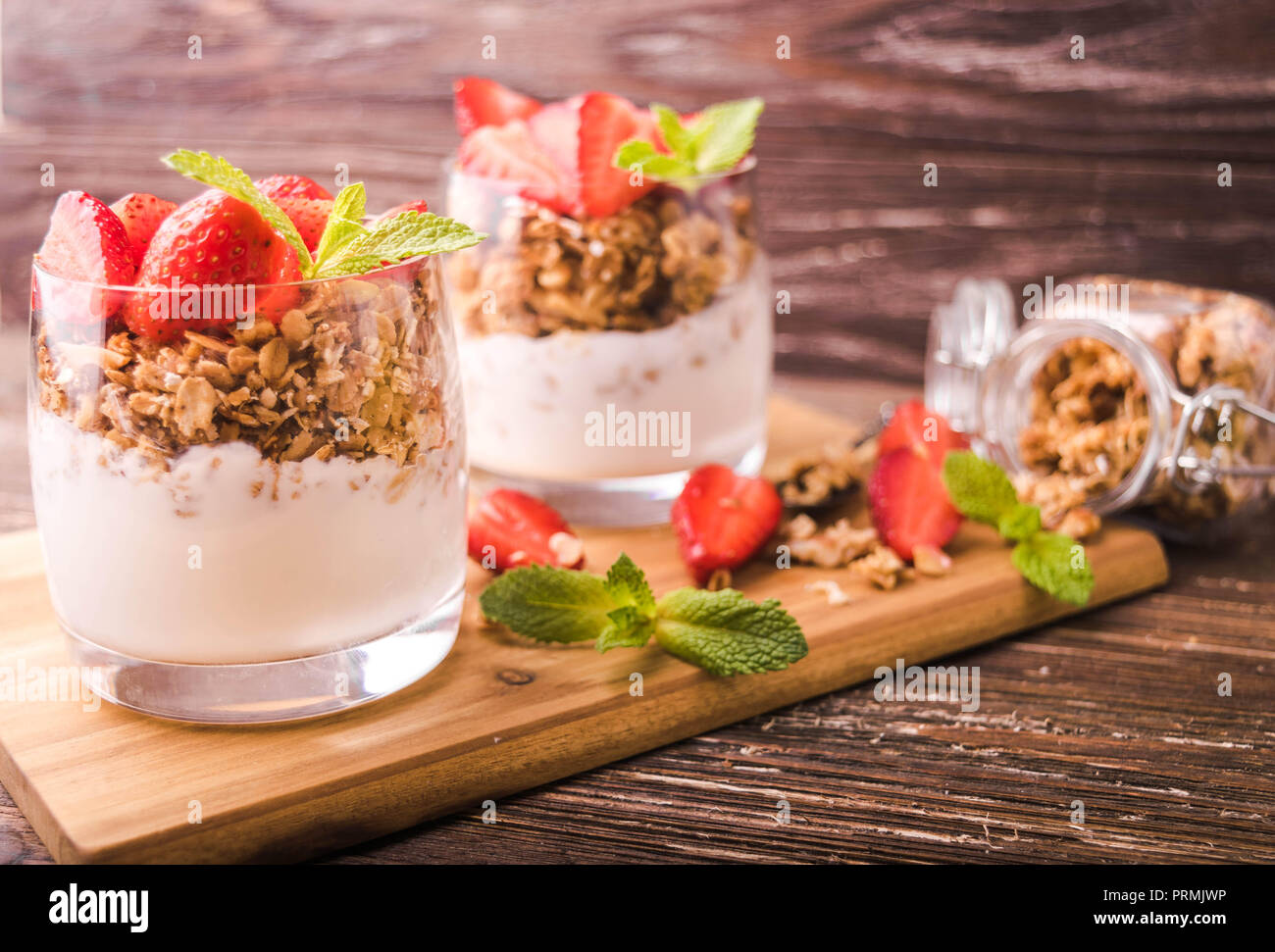 Yogurt parfait in glasses with granola, strawberries and mint leaves. On wooden board. Healthy breakfast concept. Rustic wooden table background. Stock Photo