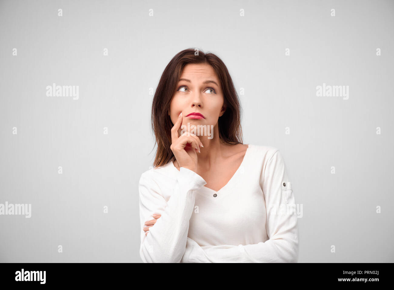 Isolated portrait of stylish young caucasian woman with dark hair touching her chin and looking up with doubtful and sceptical expression, suspecting  Stock Photo