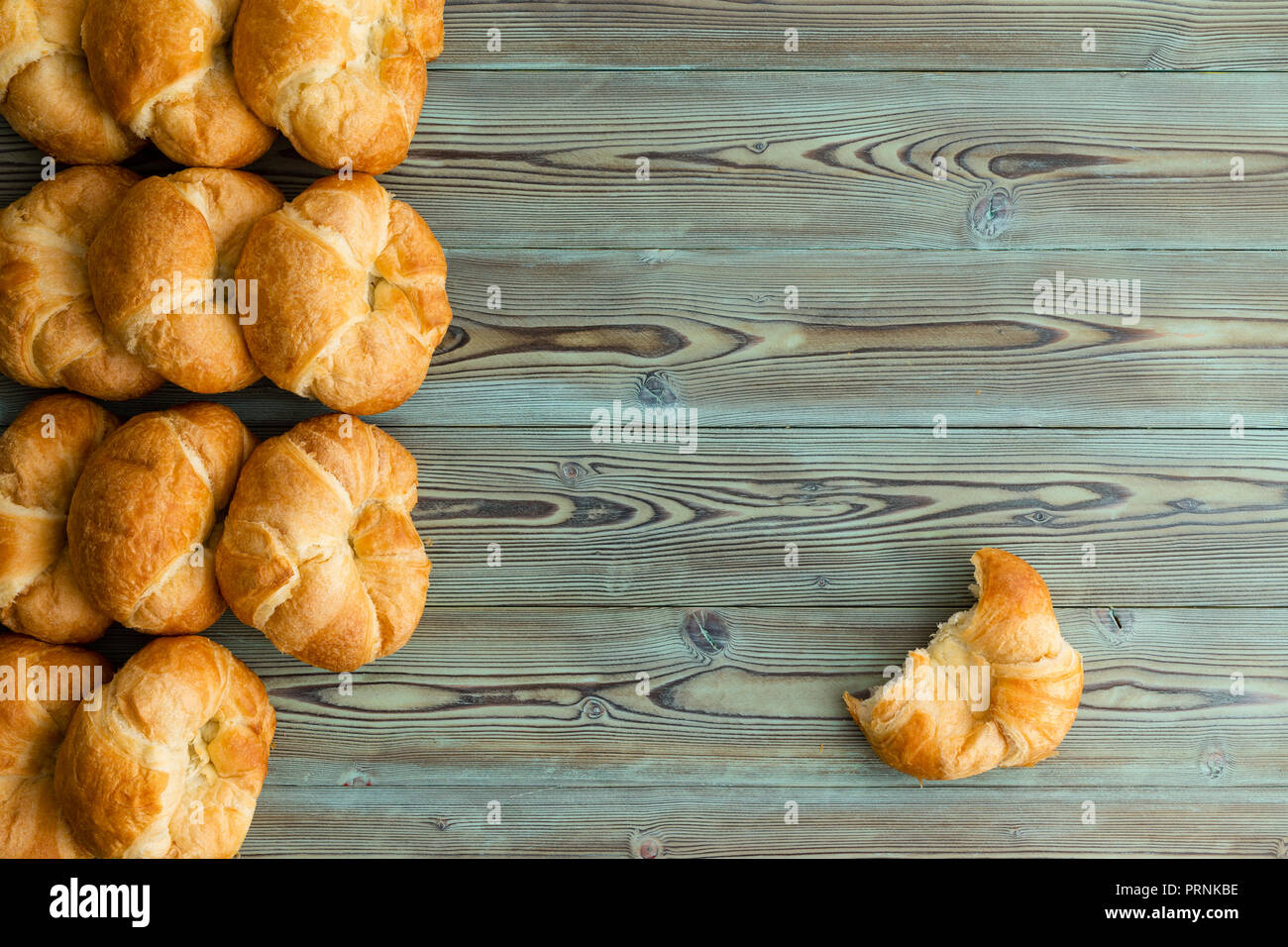 Rows of freshly baked croissants in a side border with a single half eaten croissant to the side on rustic stained wood with copy space Stock Photo