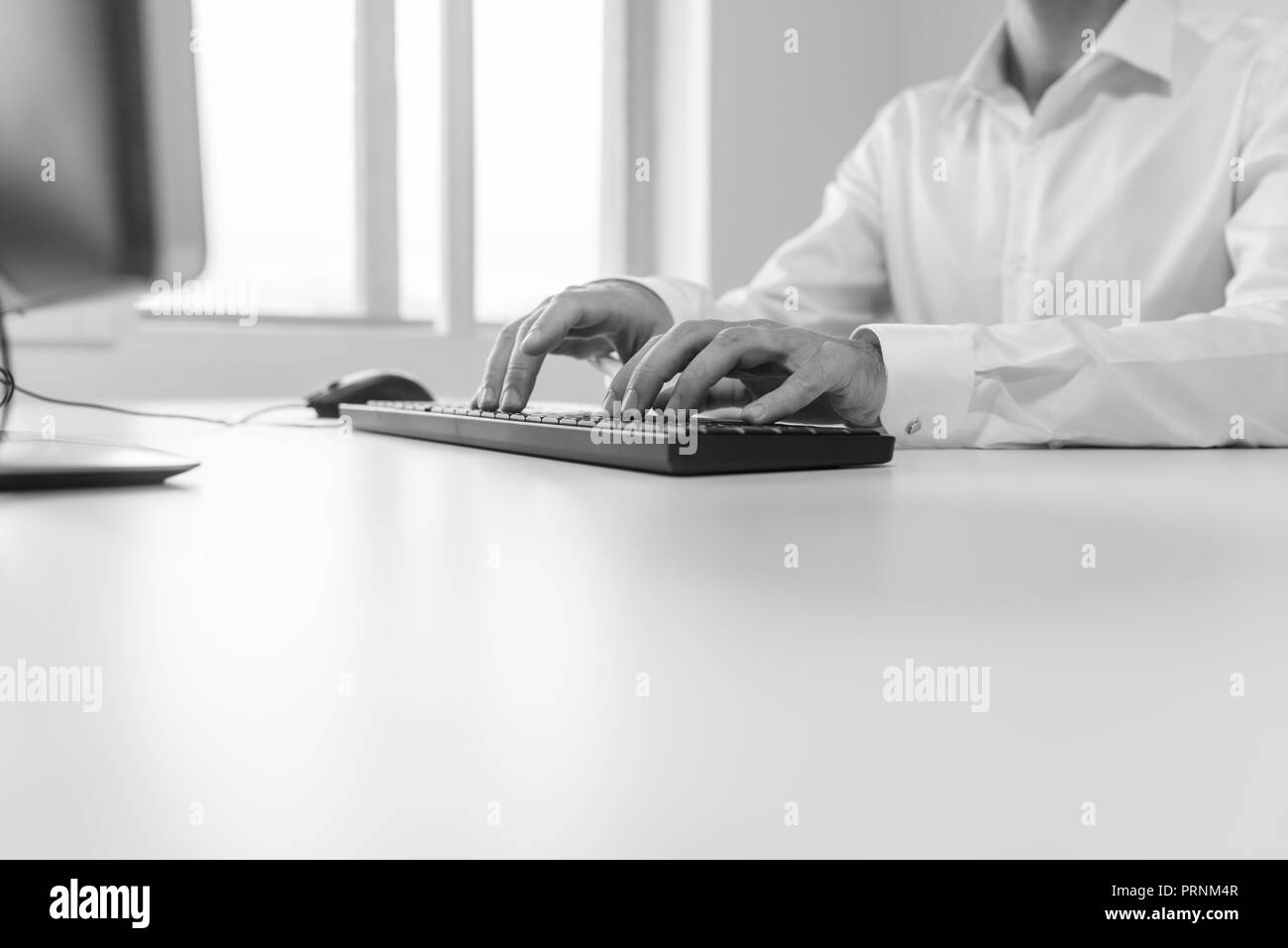 Black and white image of businessman sitting at his office desk typing on computer keyboard. Stock Photo