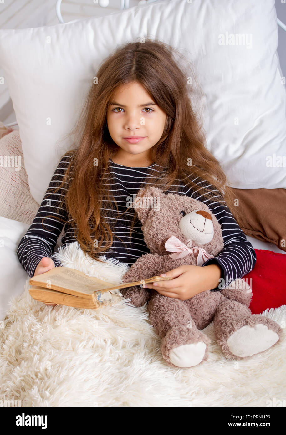 Little girl lying on the bed with her favorite teddy bear and reading a book before bedtime Stock Photo