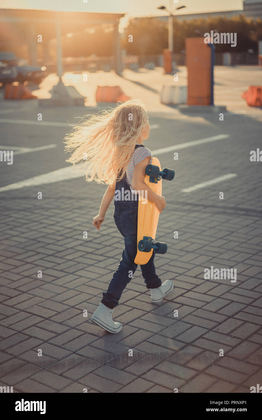 rear view of child walking with skateboard in hands at parking lot ...