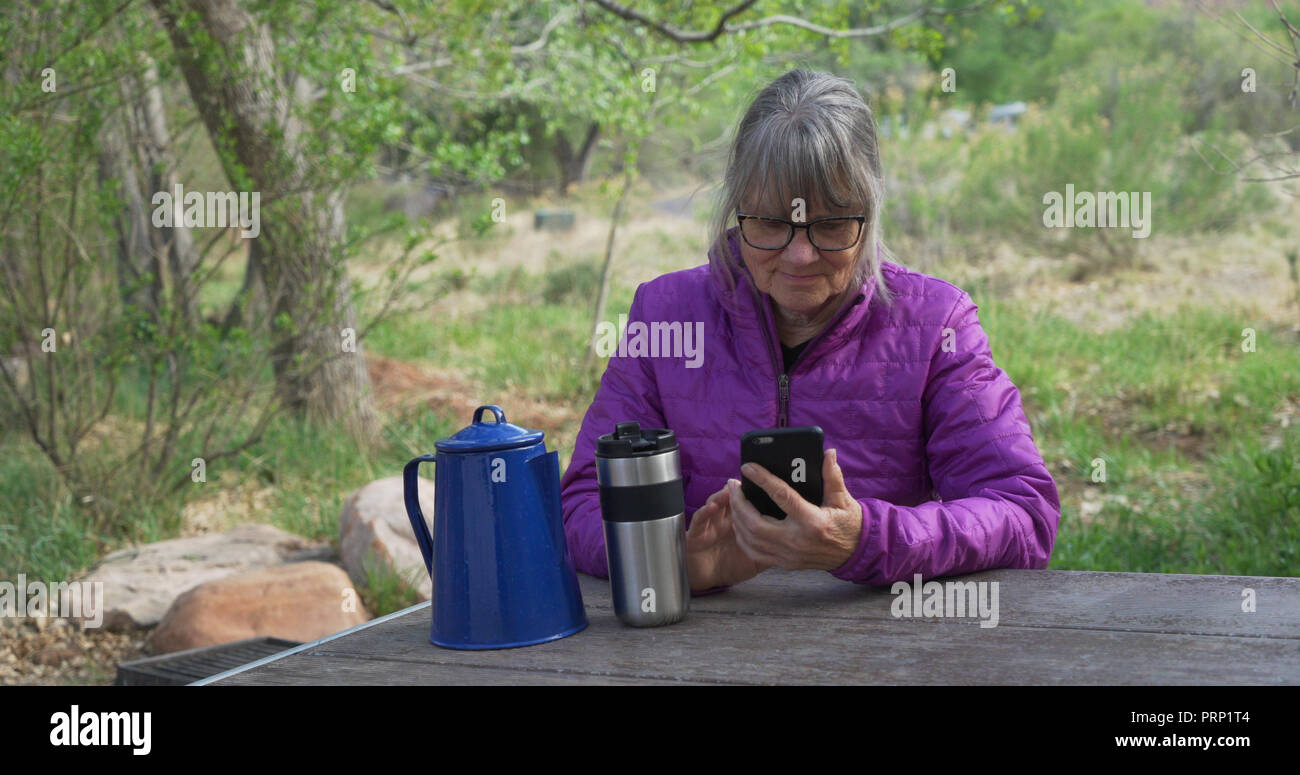 Elderly female hiker resting on bench reading smartphone outdoors in nature Stock Photo