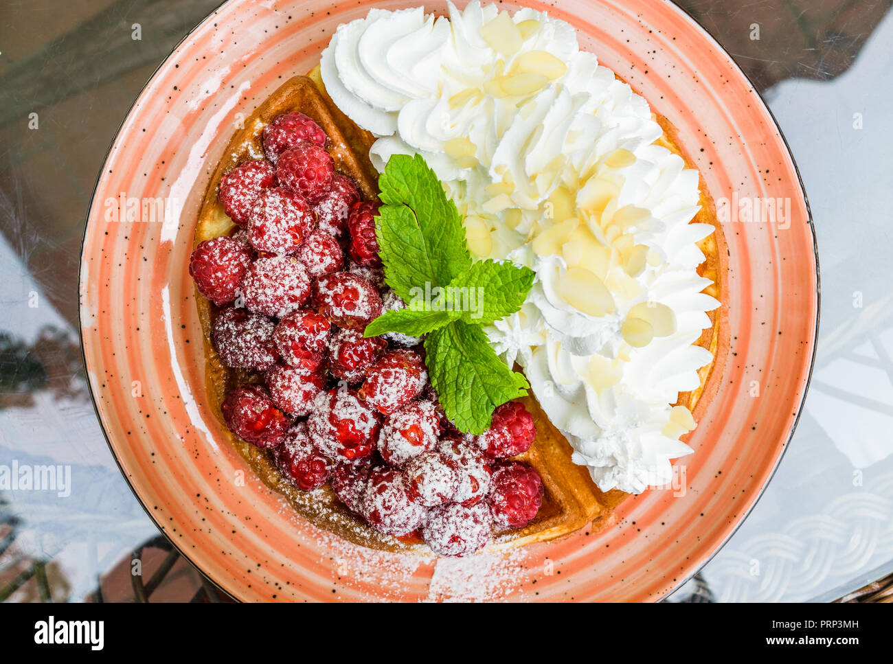 Tasty belgian waffles with fresh raspberries and whipped cream with almond flakes. Decorated with mint leaves and powdered sugar, on orange plate. Del Stock Photo