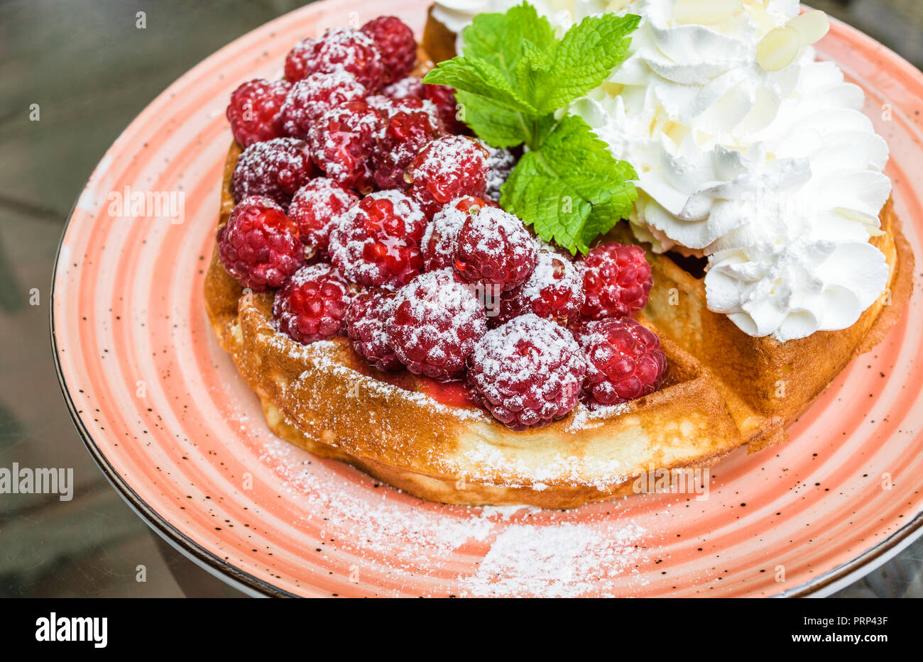 Tasty belgian waffles with fresh raspberries and whipped cream with almond flakes. Decorated with mint leaves and powdered sugar, on orange plate. Del Stock Photo