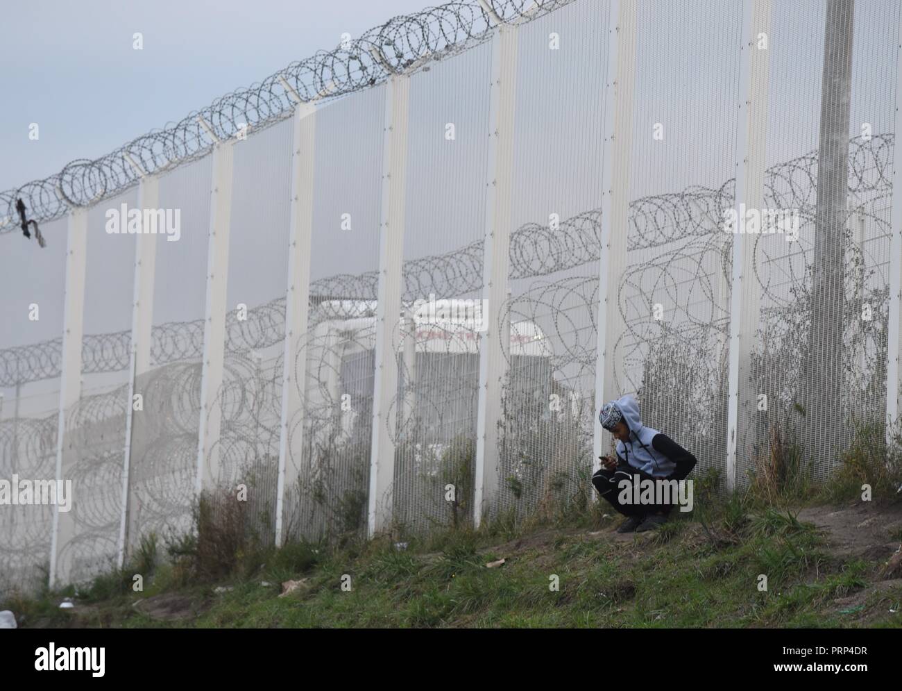 October 12, 2016 - Calais, France: A migrant using his mobile phone near the high-security fences erected between the Calais 'jungle' migrant camp and the road leading to the port. Most migrants living in this camp attempt to illegally enter the United Kingdom. Scene de vie quotidienne dans la jungle de Calais, l'un des plus grands camps de migrants au monde. *** FRANCE OUT / NO SALES TO FRENCH MEDIA *** Stock Photo