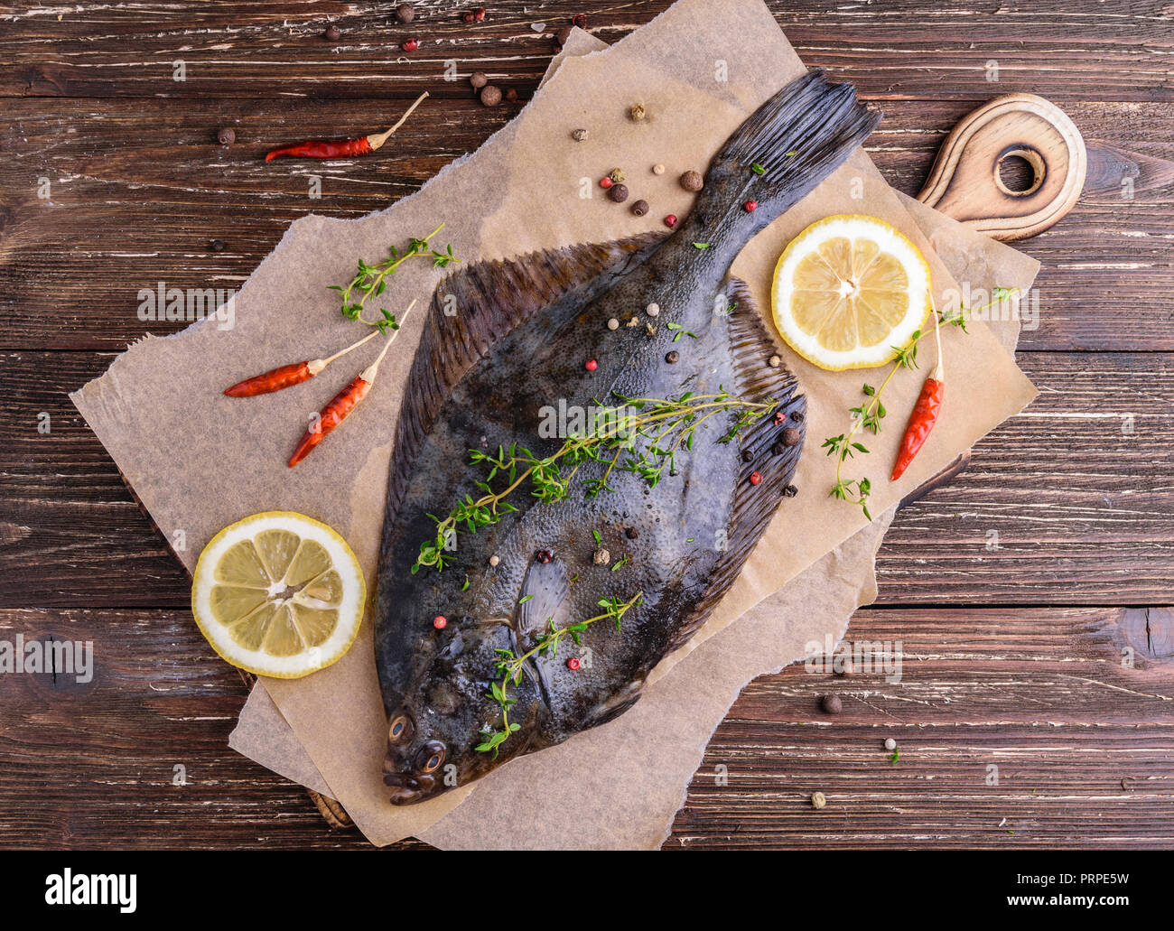 Raw flounder plaice fish (flatfish). Cooking process concept. Flounder fish with spices, lemon slices, thyme on parchment paper. Dark wooden table bac Stock Photo