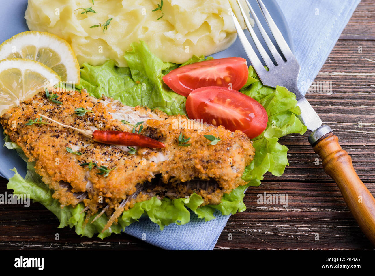 Fried flat fish on green lettuce with mashed potatoes, tomato, pepper and lemon slices in blue plate on dark rustic wooden background. Delicious dinne Stock Photo