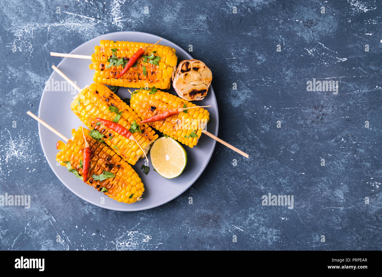Grilled corn cobs. Delicious summer snack. With parsley, chili peppers, lime and grilled garlic. Served on blue plate. Dark blue stone concrete backgr Stock Photo
