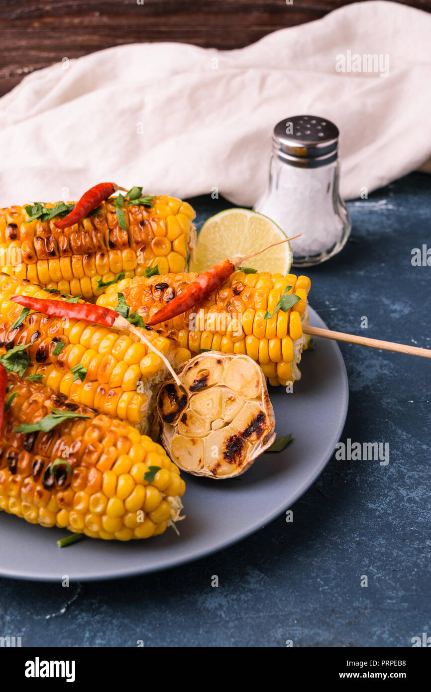 Grilled corn cobs. Delicious summer snack. With parsley, chili peppers, lime, salt and grilled garlic. Served on blue plate. Dark blue stone concrete  Stock Photo