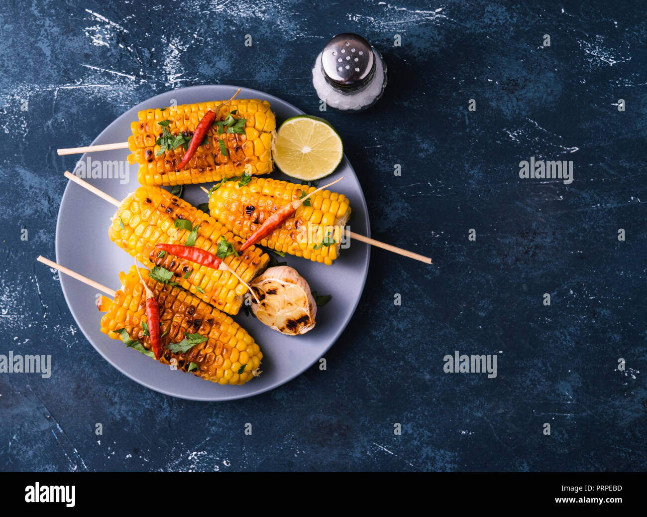 Grilled corn cobs. Delicious summer snack. With parsley, chili peppers, lime and grilled garlic. Served on blue plate. Dark blue stone concrete backgr Stock Photo