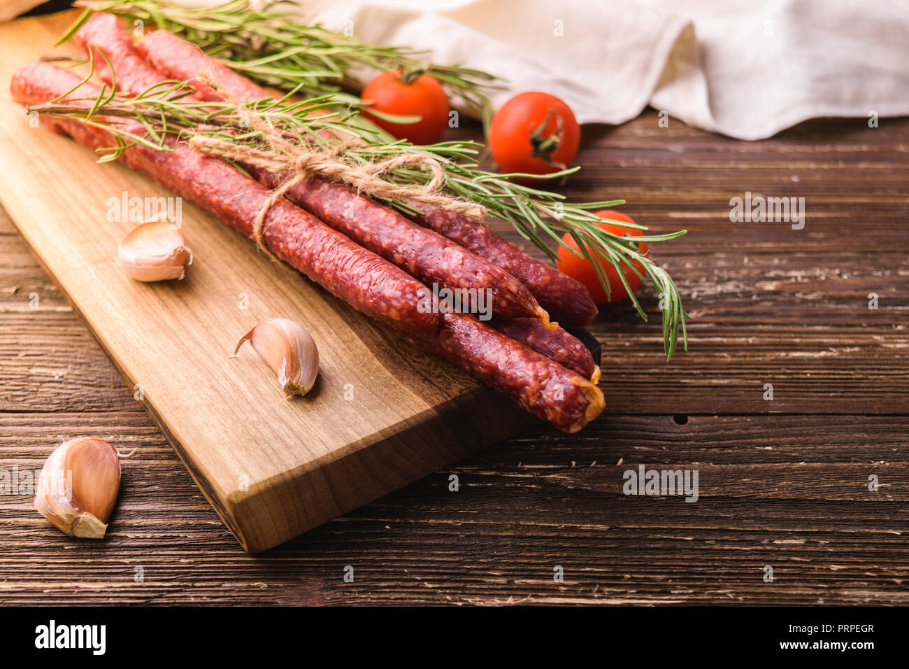 Kabanosy - smoked traditional polish pork sausages on wooden cutting board. With rosemary herb, garlic cloves and tomatoes. On rustic wooden table Stock Photo