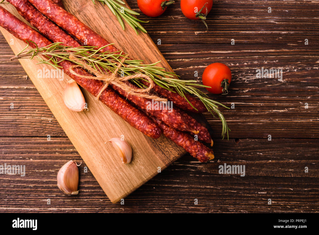 Kabanosy - smoked traditional polish pork sausages on wooden cutting board. With rosemary herb, garlic cloves and tomatoes. On rustic wooden table. To Stock Photo