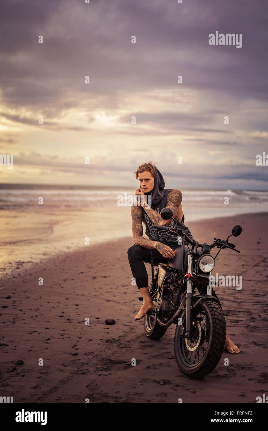 handsome tattooed biker posing on motorcycle on ocean beach Stock Photo