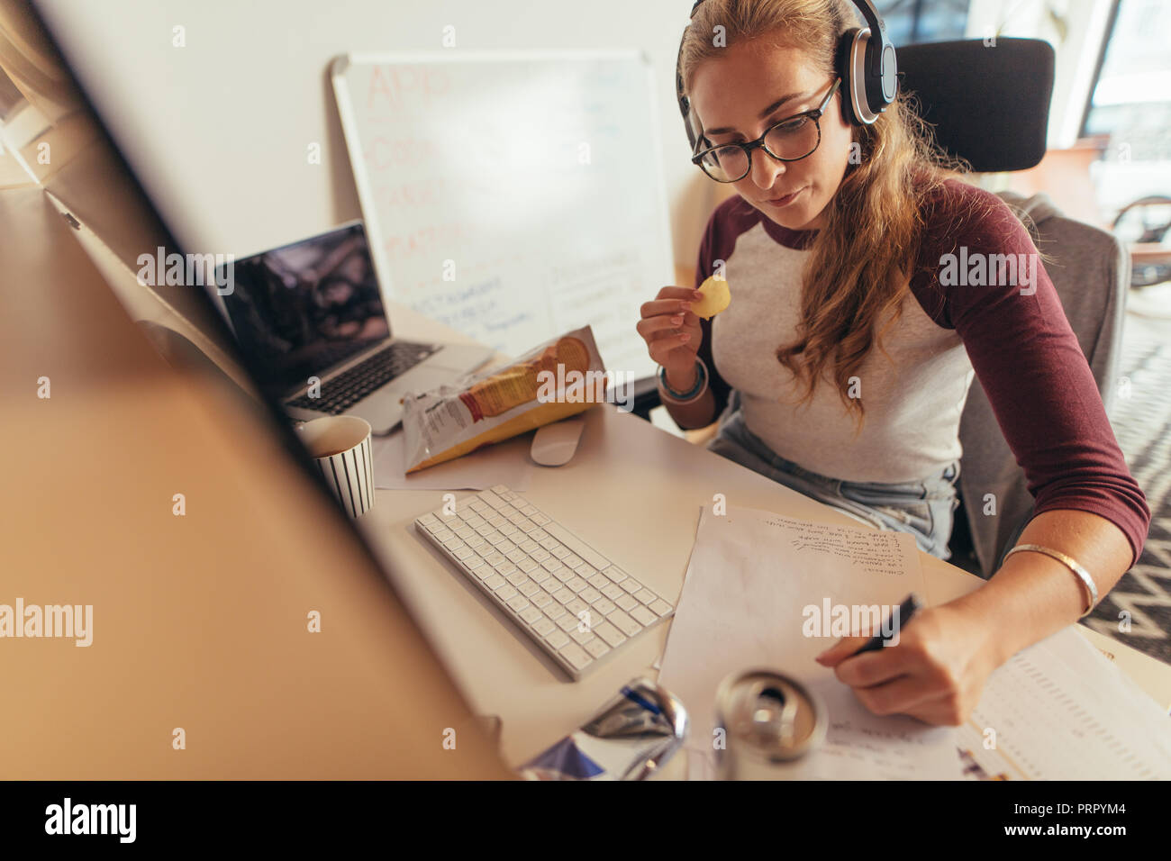 Woman writing programme codes on paper and eating. Female coder working on new software development at tech startup in office. Stock Photo