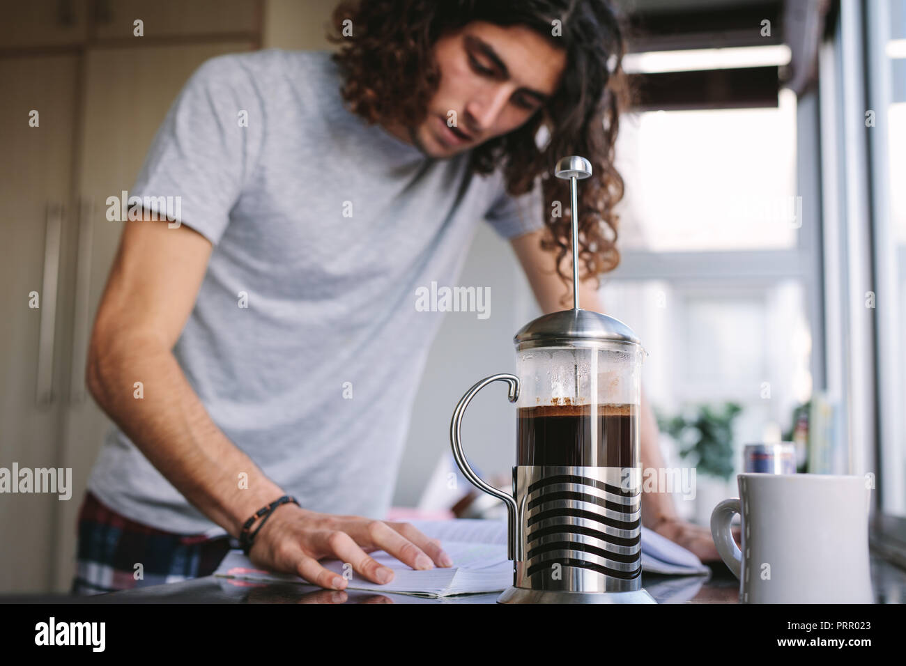 Young man reading book standing in kitchen at home while making coffee. Student standing in kitchen and studying for college exam. Stock Photo