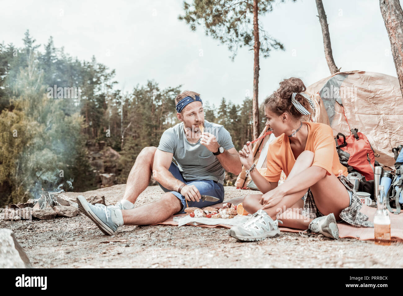 Two happy backpackers trying delicious grilled sausages while sitting near tent Stock Photo