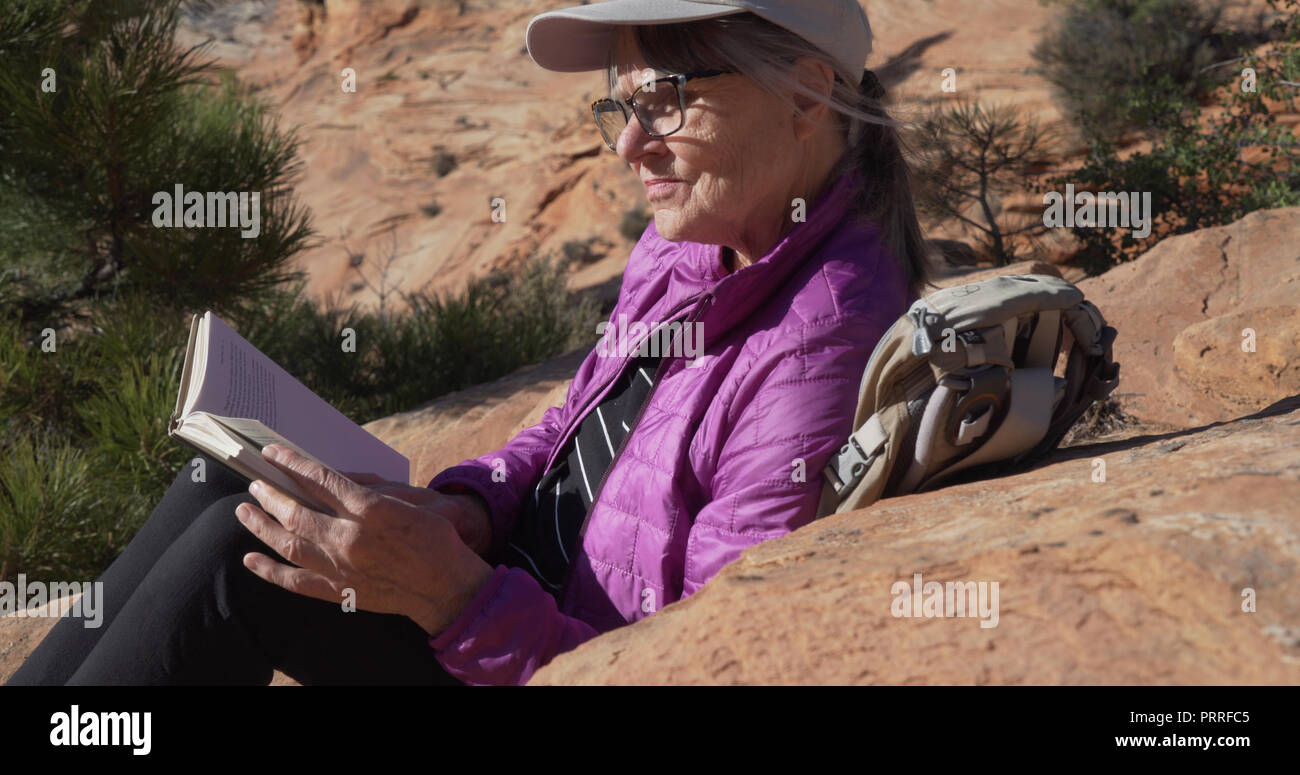 Close up portrait of elderly woman resting on rocky surface reading a novel Stock Photo