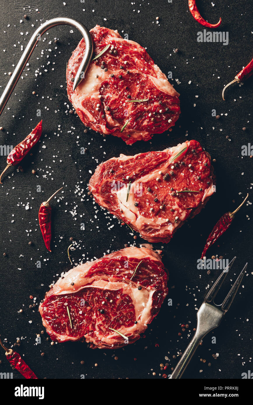 top view of raw steaks, scattered spices, meat fork and metal hook on table in kitchen Stock Photo