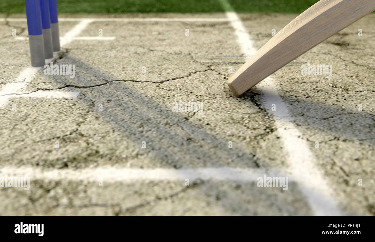 A tip of a cricket bat sliding over the crease on a cracked grass pitch