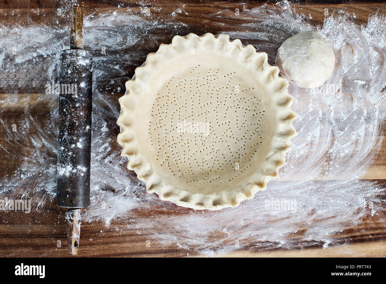 Homemade butter pie crust in pie plate with fluted pinched edge, rolling pin and extra ball of dough over floured rustic wooden background. Crust has  Stock Photo
