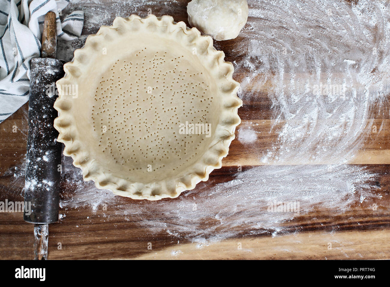 Homemade butter pie crust in pie plate with fluted pinched edge, rolling pin, towel and extra ball of dough over floured rustic wooden background. Stock Photo