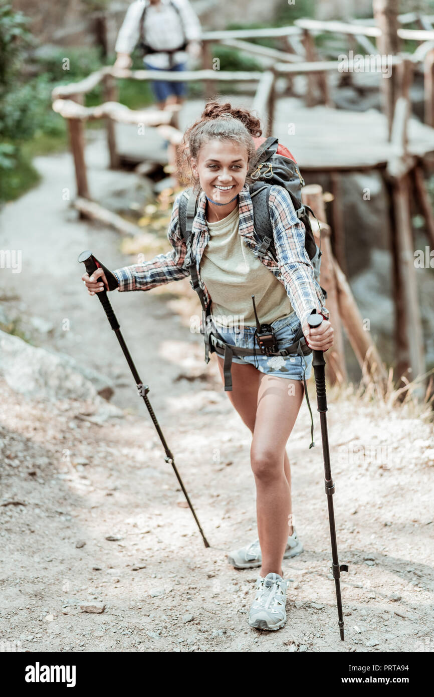 Beaming dark-haired woman feeling amazing while hiking with walking poles Stock Photo
