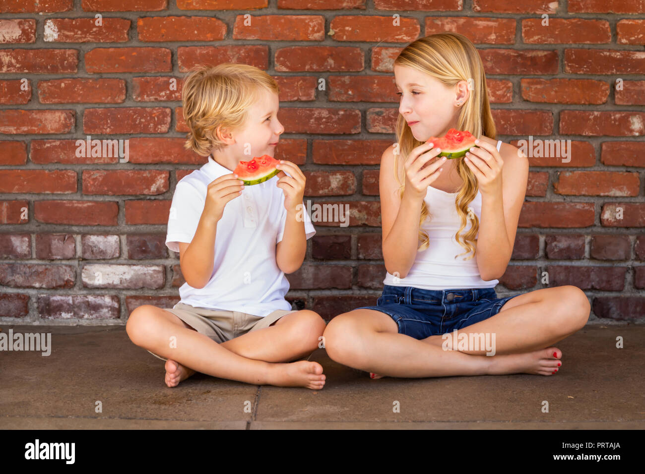 Cute Young Cuacasian Boy and Girl Eating Watermelon Against Brick Wall. Stock Photo