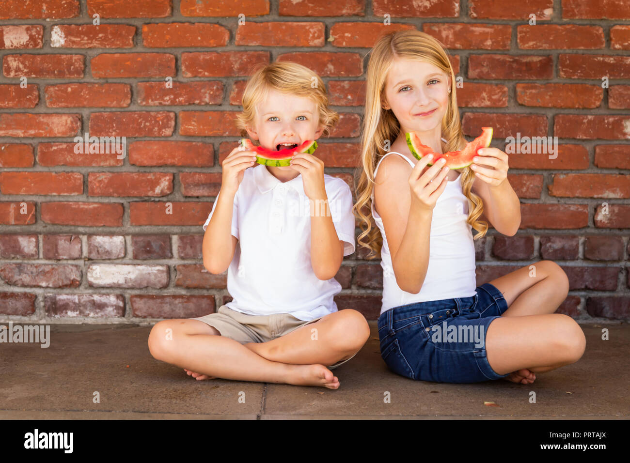 Cute Young Cuacasian Boy and Girl Eating Watermelon Against Brick Wall. Stock Photo