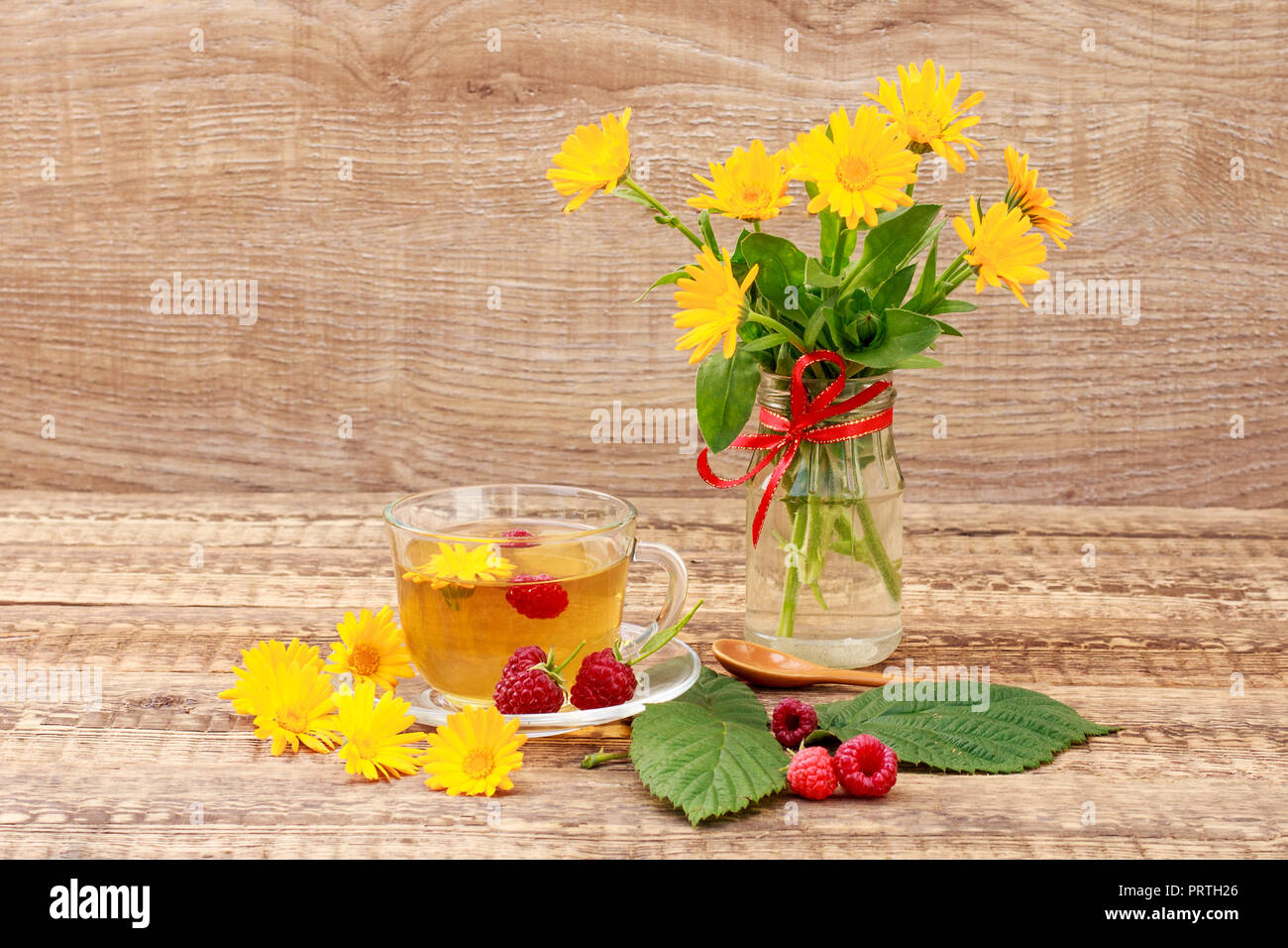 Glass cup of green tea with flowers of calendula and fresh raspberries, fresh bouquet of orange calendula in vase. Wooden boards background. Health-gi Stock Photo