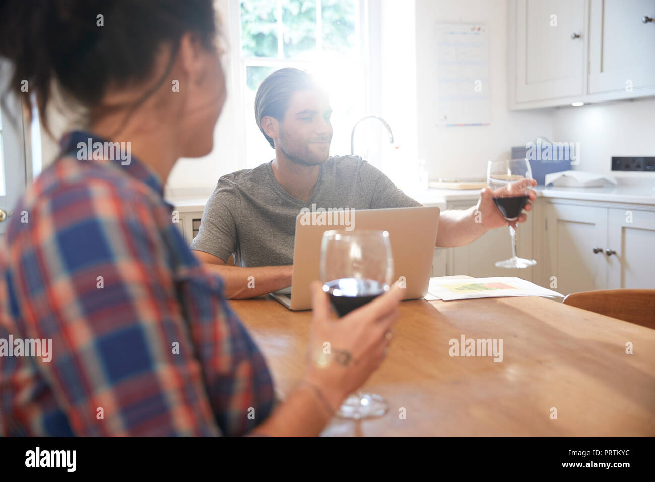 Couple at kitchen table drinking red wine while using laptop Stock Photo