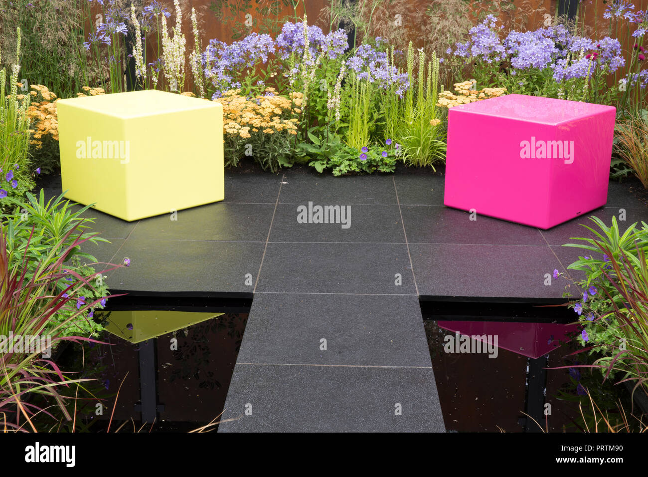 Modern Pink and yellow plastic cube box seat dark modern stone paving paved patio area small dark reflective pond colourful mixed flower borders Stock Photo
