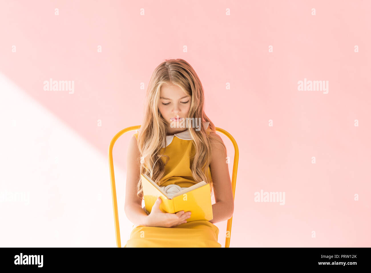 beautiful blonde youngster reading book while sitting on yellow chair on pink Stock Photo