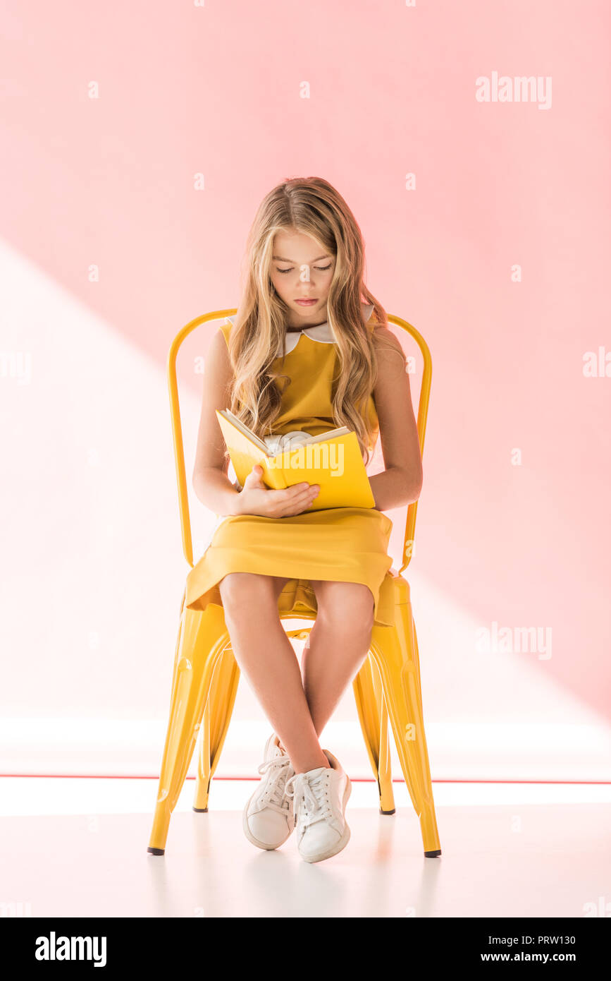 charming female youngster reading book while sitting on yellow chair on pink Stock Photo