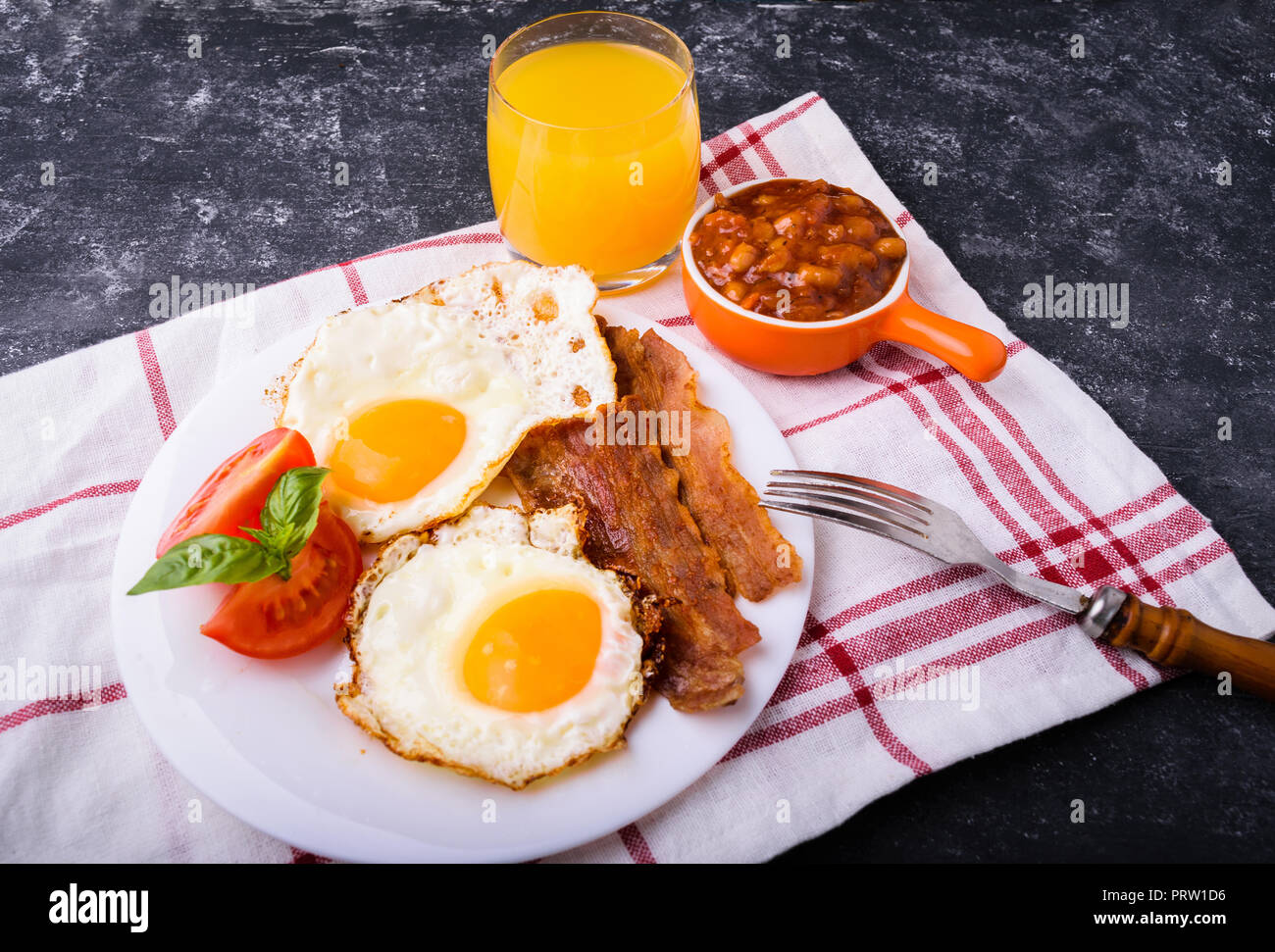English breakfast - bacon, fried eggs, beans and orange juice. Served on white plate with sliced tomato and basil leaves. Dark grunge concrete table b Stock Photo