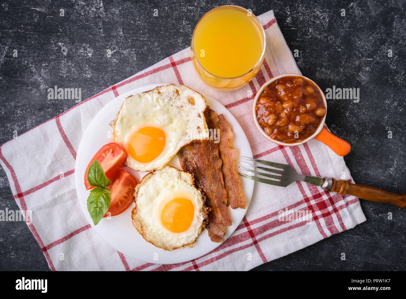 English breakfast - bacon, fried eggs, beans and orange juice. Served on white plate with sliced tomato and basil leaves. Dark grunge concrete table b Stock Photo