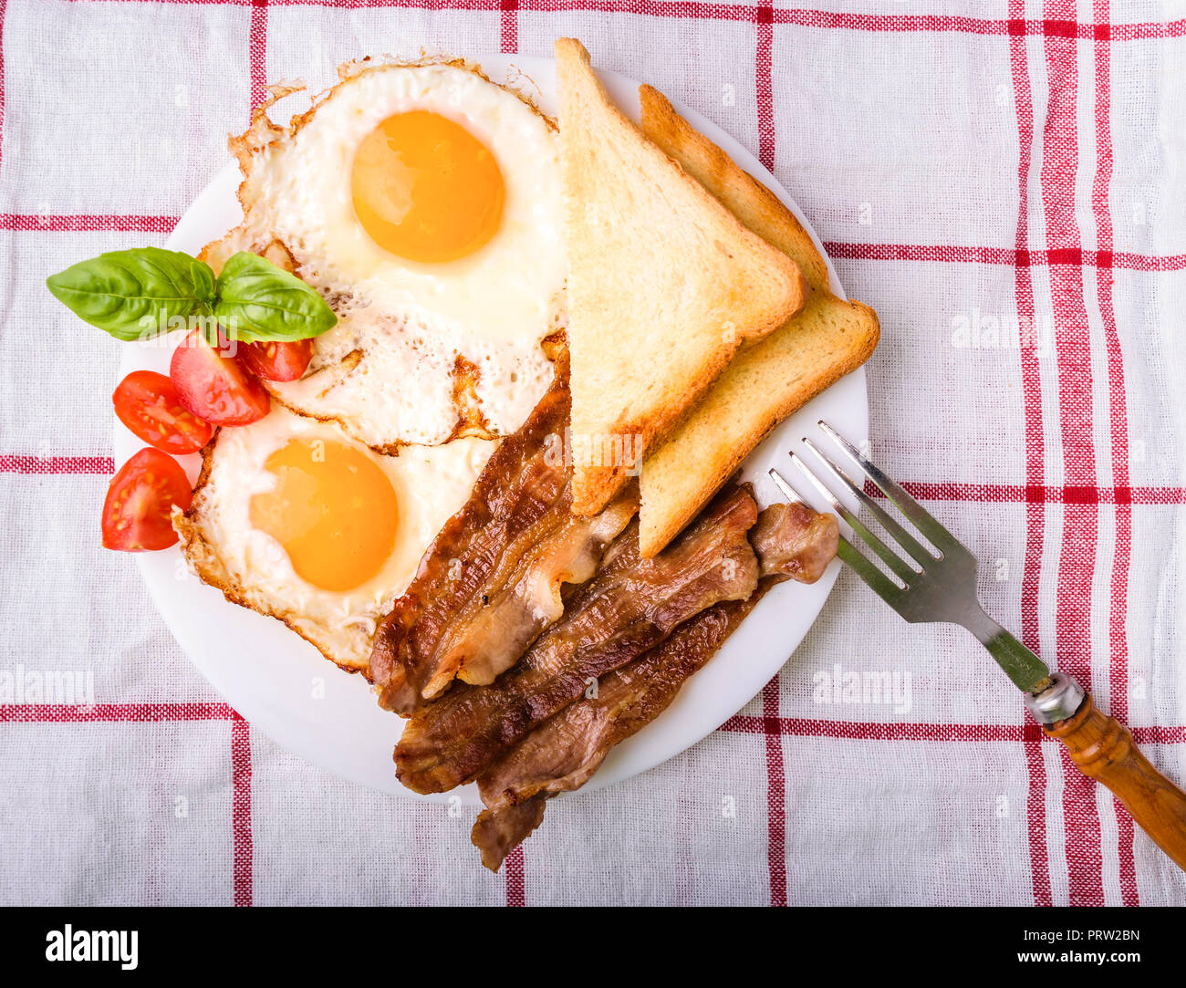 Breakfast with bacon and fried eggs. Served on white plate with sliced cherry tomato, basil leaves and crispy toast slice. Top view, view from above. Stock Photo