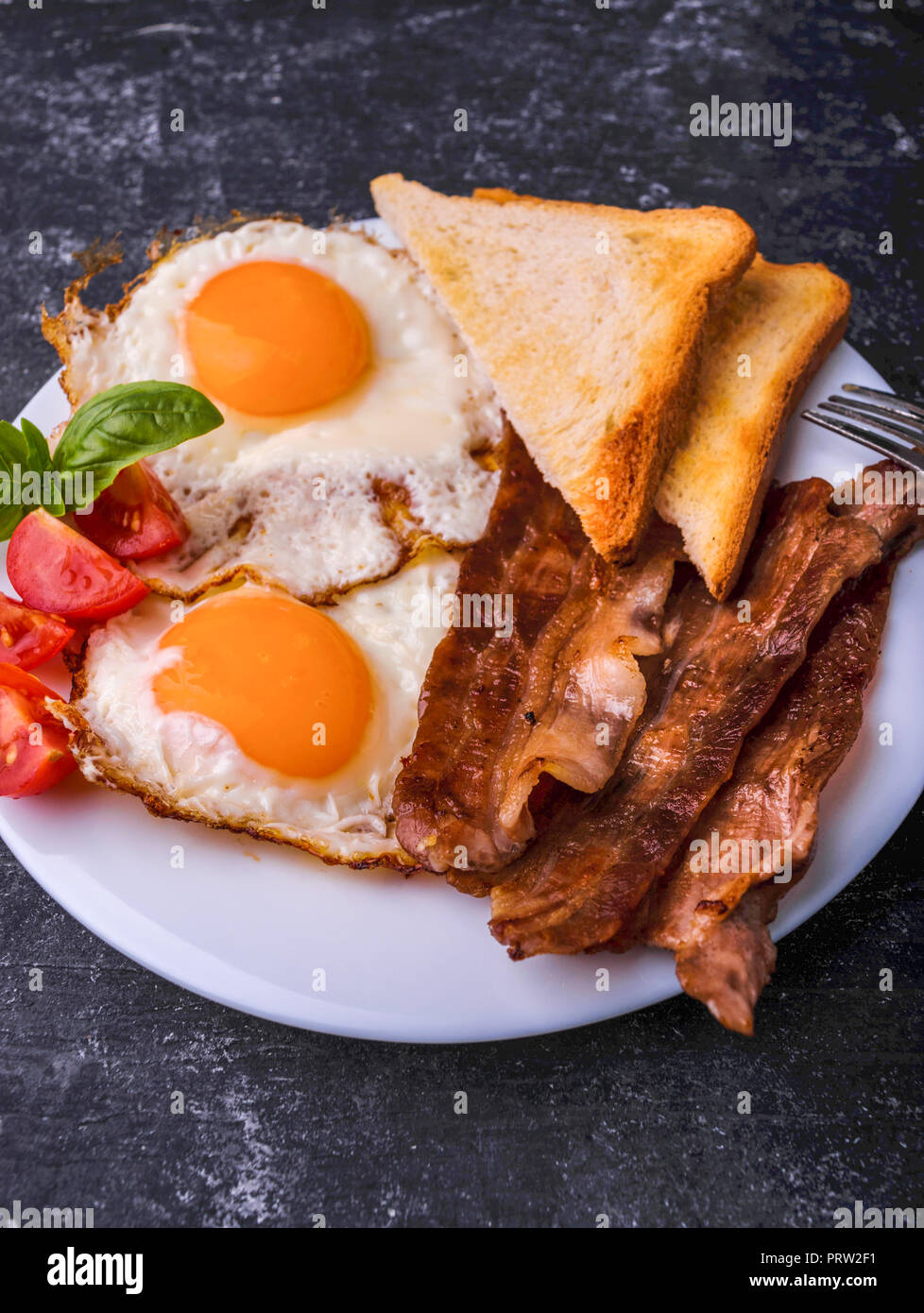 Breakfast with bacon and fried eggs. Served on white plate with sliced cherry tomato, basil leaves and crispy toast slice. Dark grunge concrete table  Stock Photo