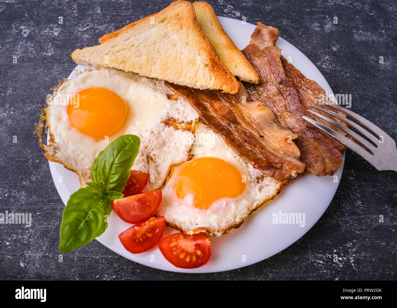 Breakfast with bacon and fried eggs. Served on white plate with sliced cherry tomato, basil leaves and crispy toast slice. Dark grunge concrete table  Stock Photo