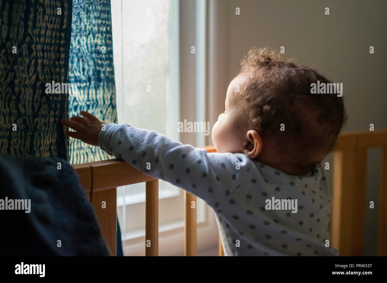 Baby boy standing up in crib looking out through window Stock Photo