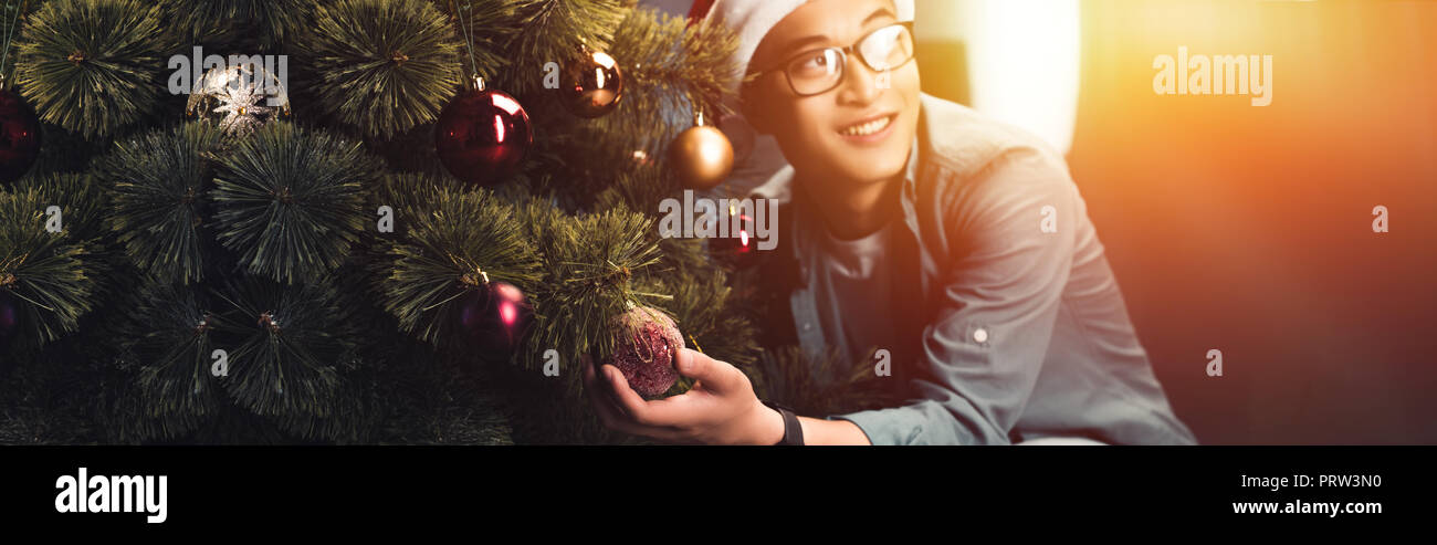 handsome happy young asian man in santa hat and eyeglasses decorating christmas tree and looking away Stock Photo