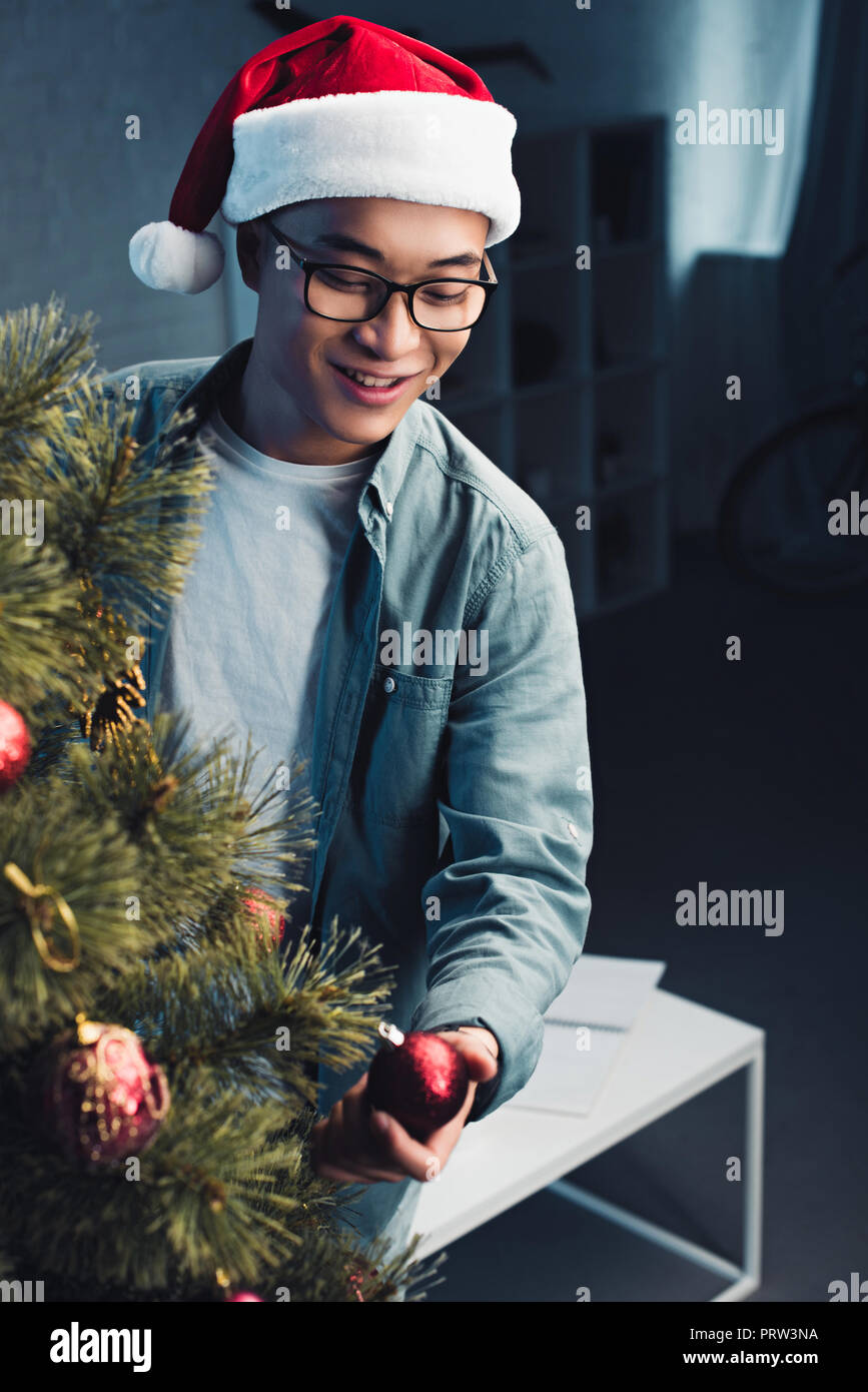 handsome smiling young asian man in santa hat decorating christmas tree at home Stock Photo